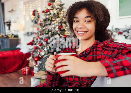 Cute African woman in pajamas sitting on couch and holding mug in bedroom. Smiling black girl is drinking coffee at home on New Stock Photo