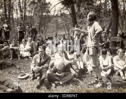 In this 1929 photograph, a robed, long-haired, guru-like figure addresses a group of men and women seated on the ground in a forest at the first Landlopers Congress in Stuttgart, Germany. The figure is gesturing while speaking. The image documents speaker-audience interaction, outdoor event organization, attire, posture, and social practices of itinerant communities in Germany in the late 1920s. Stock Photo