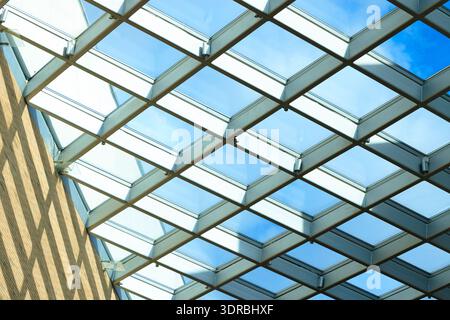 A contemporary building's interior features a striking glass roof structure with repeating diamond shapes, revealing a clear blue sky above Stock Photo