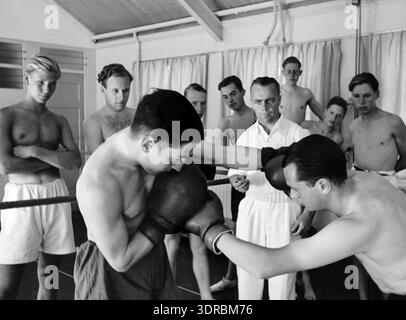 In the Netherlands on 5 December 1940, a group of men receives boxing instruction inside a sports gym. Two men are actively sparring while an instructor provides guidance. Other men stand nearby observing the session. The scene reflects sports training practices in the Netherlands during the early 20th century, emphasizing boxing techniques, physical fitness, and instructional methods in a structured gym environment. Stock Photo