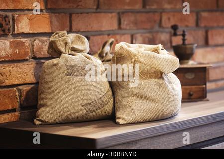 Two burlap sacks on wooden table against brick wall. Rustic storage of grains or flour in natural fabric bags in warm vintage kitchen interior Stock Photo