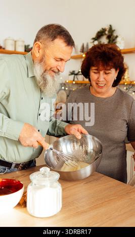 A middle-aged couple are laughing and cooking together in the kitchen at home. A mature man with a beard whips eggs for dough wi Stock Photo