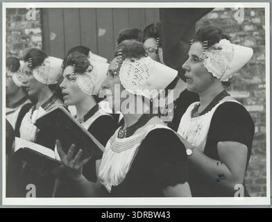 On July 28, 1947, at a camp for foreign students in West-Capelle, Netherlands, guests are being serenaded by a women's choir dressed in traditional Zeeland costume. The photograph highlights cultural exchange, traditional clothing, music performance, group singing, and the social gathering at an educational camp, emphasizing post-war cultural activities and regional folklore, as well as international student engagement and organized outdoor events in West-Capelle. Stock Photo