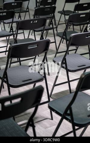 Rows of empty black folding chairs in a bright room. Empty black plastic chairs stand in rows on a light laminate floor in a classroom. Stock Photo