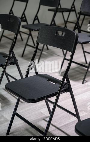 Rows of empty black folding chairs in a bright room. Empty black plastic chairs stand in rows on a light laminate floor in a classroom. Stock Photo