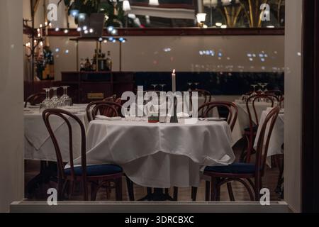Cozy restaurant interior with empty tables covered in white tablecloths, set with wine glasses and candles. Night city reflections visible in the wind Stock Photo
