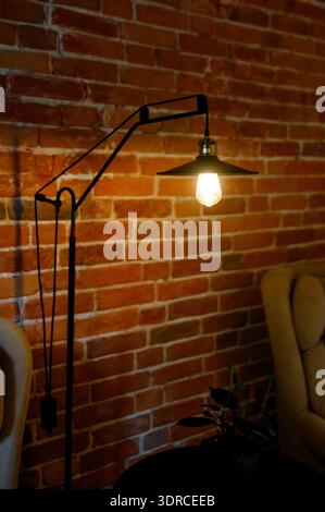A black floor lamp with an Edison bulb illuminates a brick wall in a loft-style interior.Metal adjustable lamp with warm light against red brick backg Stock Photo