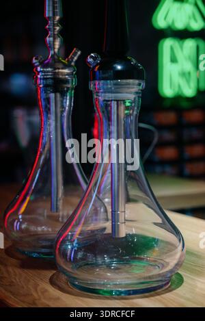 Two modern glass hookahs on a wooden stand in a dark interior with neon lighting.Professional hookahs assembled against a brick wall and green neon in Stock Photo