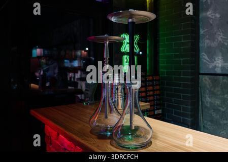 Two modern glass hookahs on a wooden stand in a dark interior with neon lighting.Professional hookahs assembled against a brick wall and green neon in Stock Photo