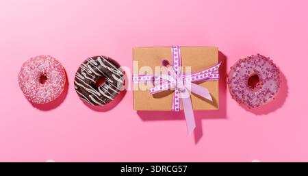A festive flat lay featuring vibrant donuts with colorful toppings and a gift box on a pink background Stock Photo