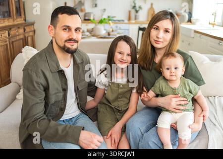 Happy family at home. Mother father two children daughters relaxing on sofa indoor. Mom dad parents baby girls kids relax playin Stock Photo