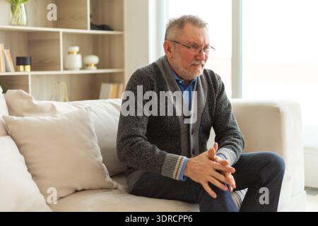 Knee pain arthritis body sick health care concept. Unhappy middle aged senior man suffering from knee ache sitting on sofa at ho Stock Photo
