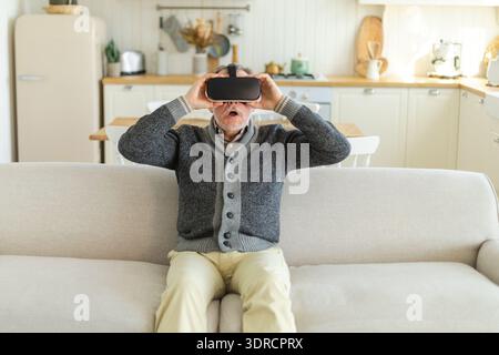 Excited mature senior man wearing using virtual reality metaverse VR glasses headset at home. Grandfather touching air during VR Stock Photo