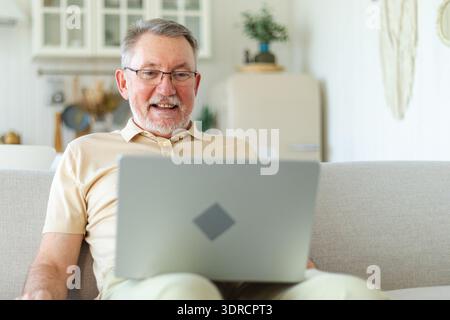 Confident stylish happy middle aged senior man using laptop at home. Stylish older mature 60s beard grandfather sitting at couch Stock Photo