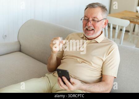 Middle aged senior man euphoric winner with smartphone. Older mature grandfather looking at cell phone reading great news gettin Stock Photo