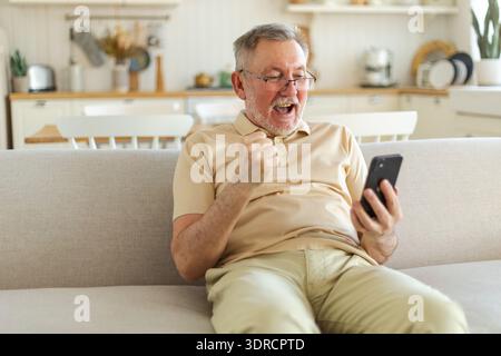 Middle aged senior man euphoric winner with smartphone. Older mature grandfather looking at cell phone reading great news gettin Stock Photo