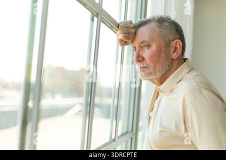 Sad tired lonely disappointed older senior man at home alone. Unhappy grandfather looking sadly outside window experiencing grie Stock Photo