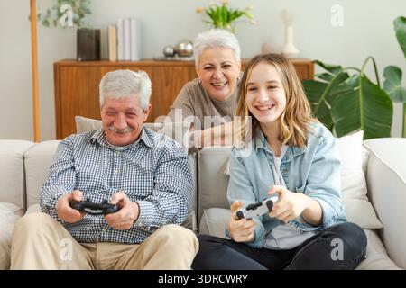 Happy family at home. Old senior man grandfather woman grandmother girl granddaughter playing video game with joysticks at home. Stock Photo