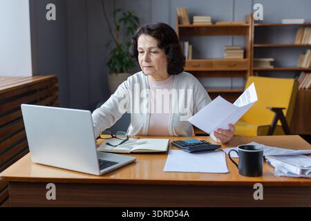 Middle aged senior woman sit with laptop and paper document, Pensive older mature lady reading paper bill pay online at home man Stock Photo
