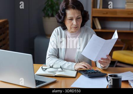 Middle aged senior woman sit with laptop and paper document, Pensive older mature lady reading paper bill pay online at home man Stock Photo