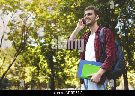 College Student Talks on Phone While Holding Notebooks in Park During Afternoon Stock Photo