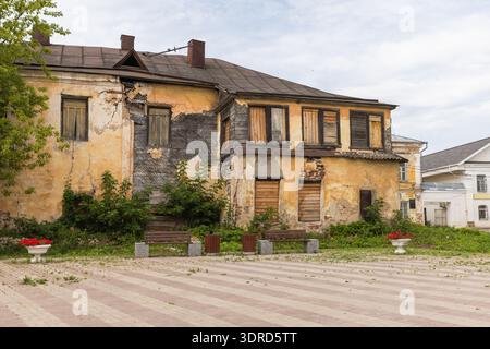 An abandoned two-story yellow houses with peeling plaster, boarded windows and visible structural damage, surrounded by overgrown greenery, benches an Stock Photo