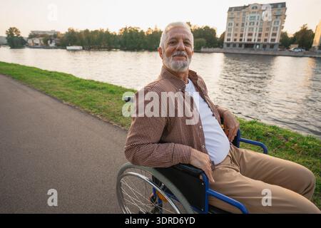Happy senior man in wheelchair smiling enjoying freedom walking on road in city park outdoor. Old cheerful positive man in chair Stock Photo