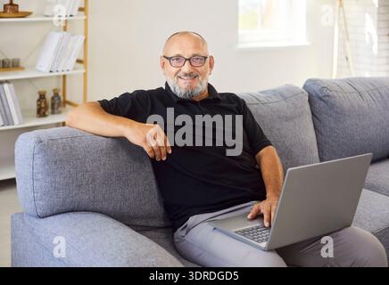 Cheerful senior man adjusting glasses while using laptop on sofa, home office mood Stock Photo