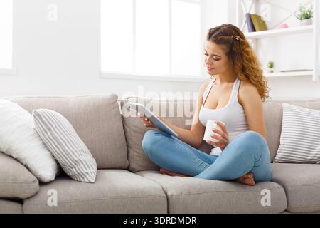 Woman Sitting on Couch Using Tablet and Holding Cup of Coffee in Living Room During Daytime Stock Photo