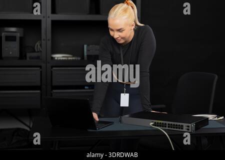 Female IT technician, young adult, blonde, working with a laptop and network switch in a tech office Stock Photo