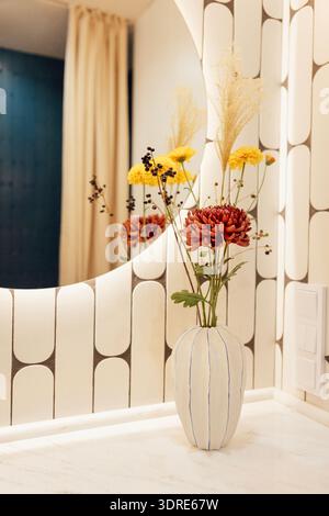 Close-up of a ceramic vase with flowers on a marble countertop in the bathroom. Stylish white tiles on the walls and a round ill Stock Photo