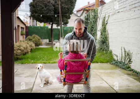 Father pushing daughter on swing in backyard with dog Stock Photo