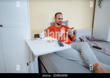 Man relaxing in bed enjoying breakfast and using smartphone Stock Photo