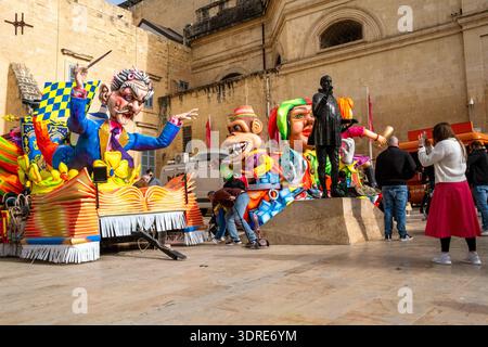 Carnival floats with oversized caricature figures are parked near a statue as visitors watch and take photos during Malta Carnival in Valletta, Malta Stock Photo