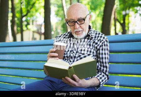 Older Man Reads a Book While Sitting on a Park Bench With a Coffee Cup in Hand Stock Photo