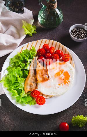 Grilled tacos with egg, fried bacon, vegetables and herbs on a plate on the table vertical view Stock Photo