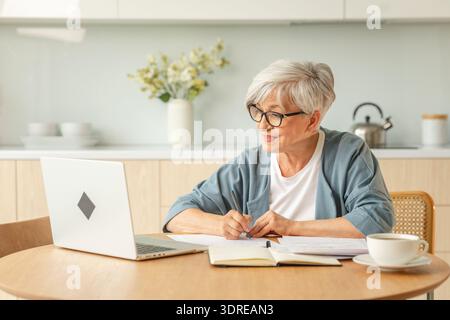Middle aged senior woman using laptop computer writing notes at home. Focused mature old businesswoman work in home office makin Stock Photo