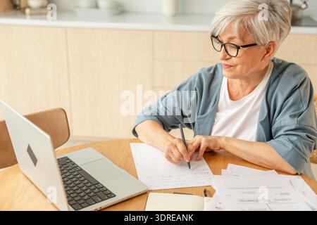 Middle aged senior woman using laptop computer writing notes at home. Focused mature old businesswoman work in home office makin Stock Photo
