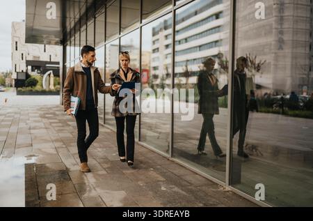 Two professionals walk together outside a modern glass building, reviewing documents Stock Photo