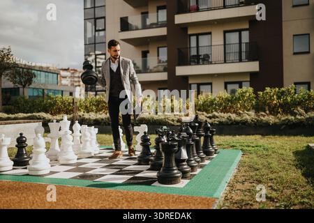 Businessman in blazer plays oversized outdoor chess near modern apartment complex Stock Photo