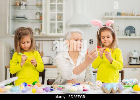 Happy Easter. Two girls sisters grandmother painting eggs having video call on phone. Happy family preparing for Easter. Family Stock Photo