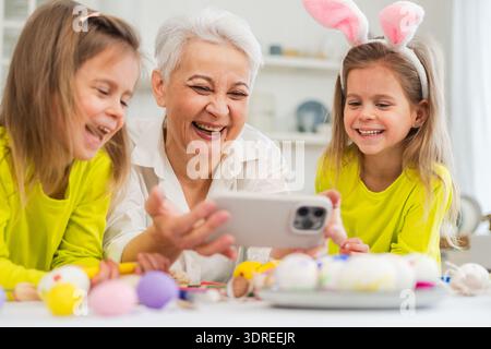 Happy Easter. Two girls sisters twins grandmother painting eggs watching video on phone enjoying time together. Happy family pre Stock Photo