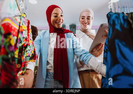 Two modern muslim women are discussing fashion options, looking at clothes on hangers while one holds a digital tablet, demonstrating shopping and sty Stock Photo