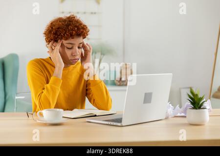 African woman touching temples experiencing stress. Girl at home office tired of working feeling headache sick rubbing temples f Stock Photo