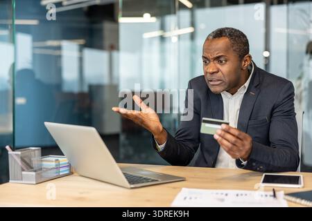 Mature businessman sitting at an office desk, holding a credit card and expressing frustration while looking at a laptop, encountering financial transaction issues during online shopping or banking Stock Photo
