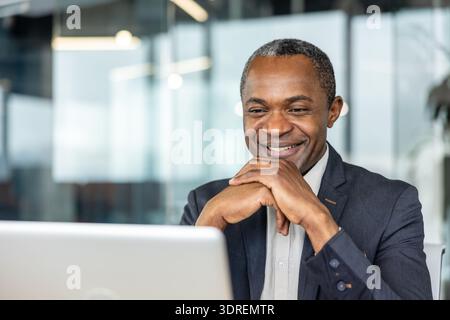 Confident male professional smiling cheerfully while looking at a laptop screen in a bright, contemporary office environment, expressing success and positive engagement Stock Photo