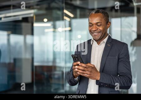 Executive man standing in a modern office building, happily using his mobile phone for professional communication and networking, representing success and technology in the contemporary workplace Stock Photo
