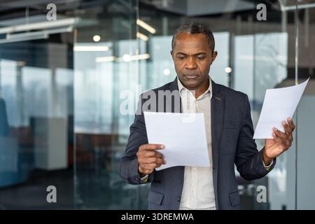 Serious african american businessman standing in a modern office, concentrating while reviewing multiple business documents and comparing information for critical decision-making Stock Photo