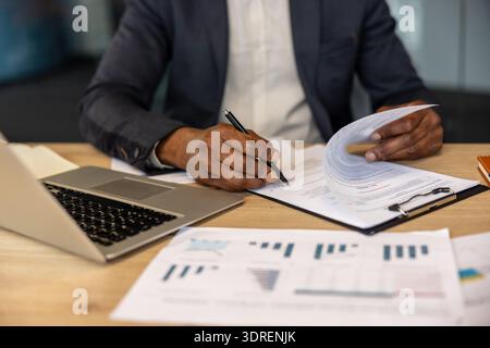 Person with dark skin tones signing a formal legal agreement on a desktop with a laptop and various white paper charts, symbolizing important corporate decisions and administrative tasks Stock Photo