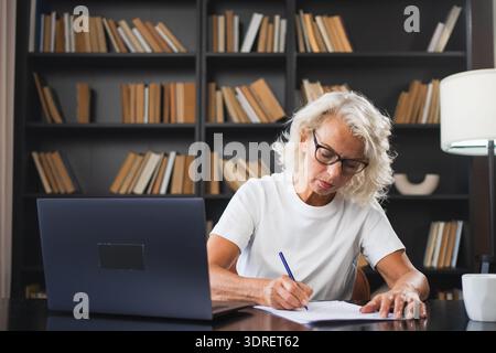 Middle aged senior woman using laptop computer writing notes at workplace. Focused mature old businesswoman work at office makin Stock Photo